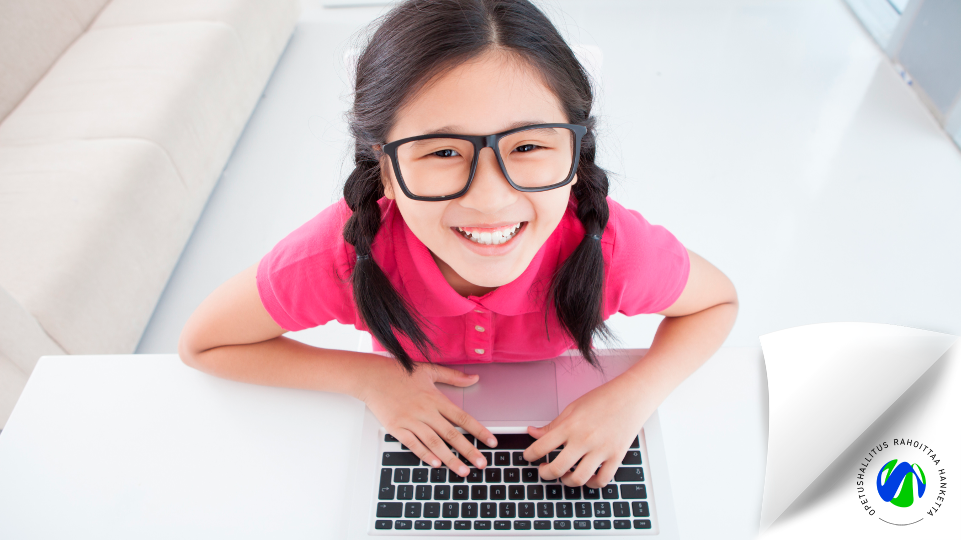 Lapsi katsoo ylöspäin kohti kameraa kädet tietokoneen näppäimistöllä ja hymyilee. OPH rahoittaa hanketta -logo alanurkassa.